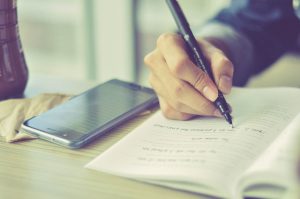 Mensch schreibt auf Notizblock, daneben liegt ein Smartphone: Digitale Medien können Hilfsmittel im Unterricht sein.