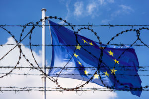 Eine Eurpaflagge weht vor blauem Himmel hinter einem Stachedrahtzaun. Das Grundrecht auf Asyl gilt in Europa! Wir müssen für Schutz der Geflüchteten sorgen, statt für Haft und Abschiebung.