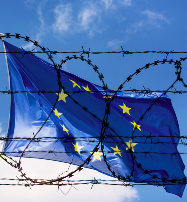 Eine Eurpaflagge weht vor blauem Himmel hinter einem Stachedrahtzaun. Das Grundrecht auf Asyl gilt in Europa! Wir müssen für Schutz der Geflüchteten sorgen, statt für Haft und Abschiebung.