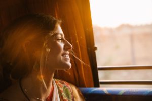 Junge lächlende Frau, blonde Haare, schaut während der Bahnfahrt aus dem Fenster, die Sonne erhellt das Bild in in warmen Farben.