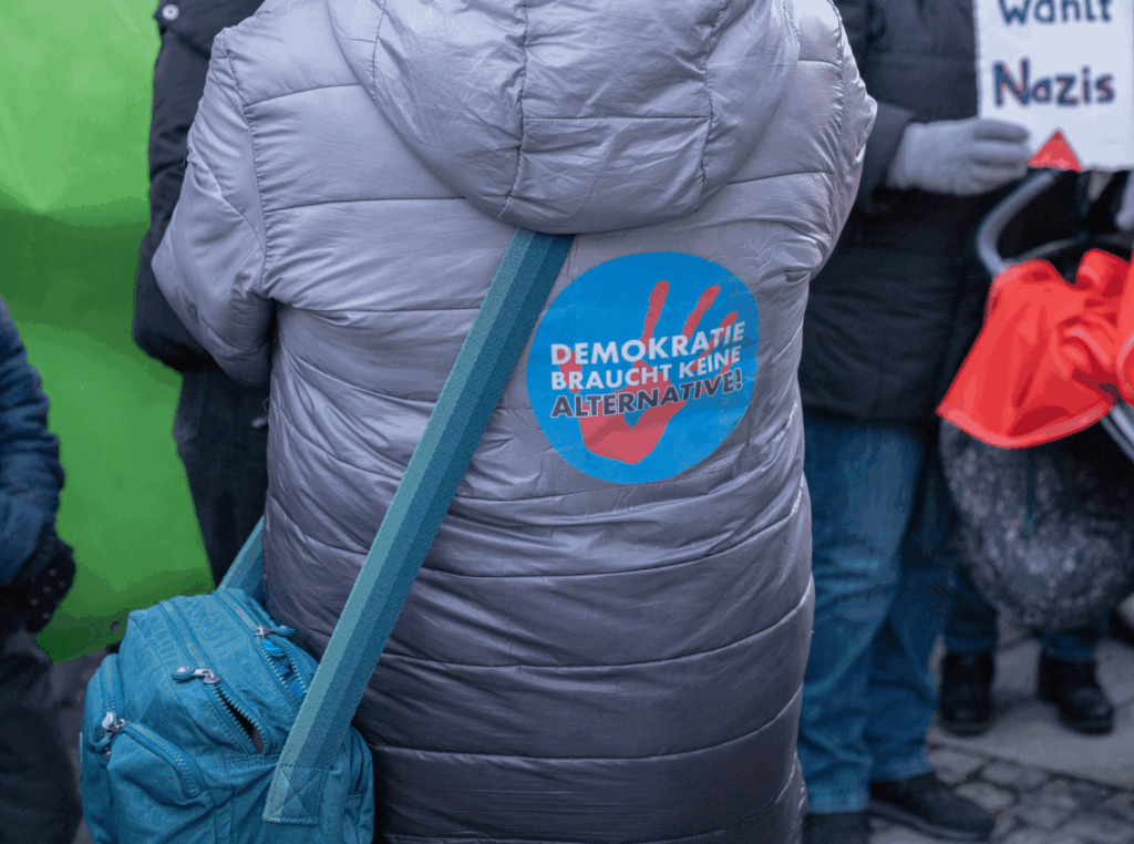 Aufkleber mit Aufschrift "Demokratie braucht keine Alternative" auf der Jackenrückseite einer Demonstrantin.