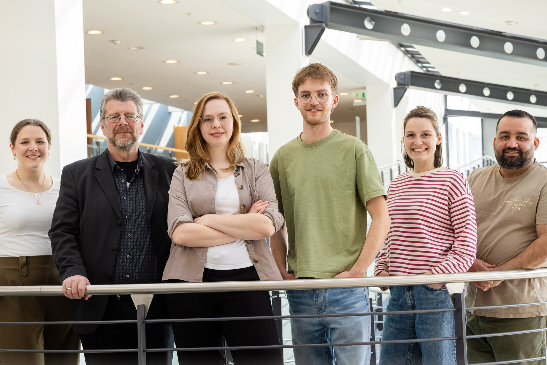 Sechs Menschen stehen an einem Geländer und lächeln in die Kamera.